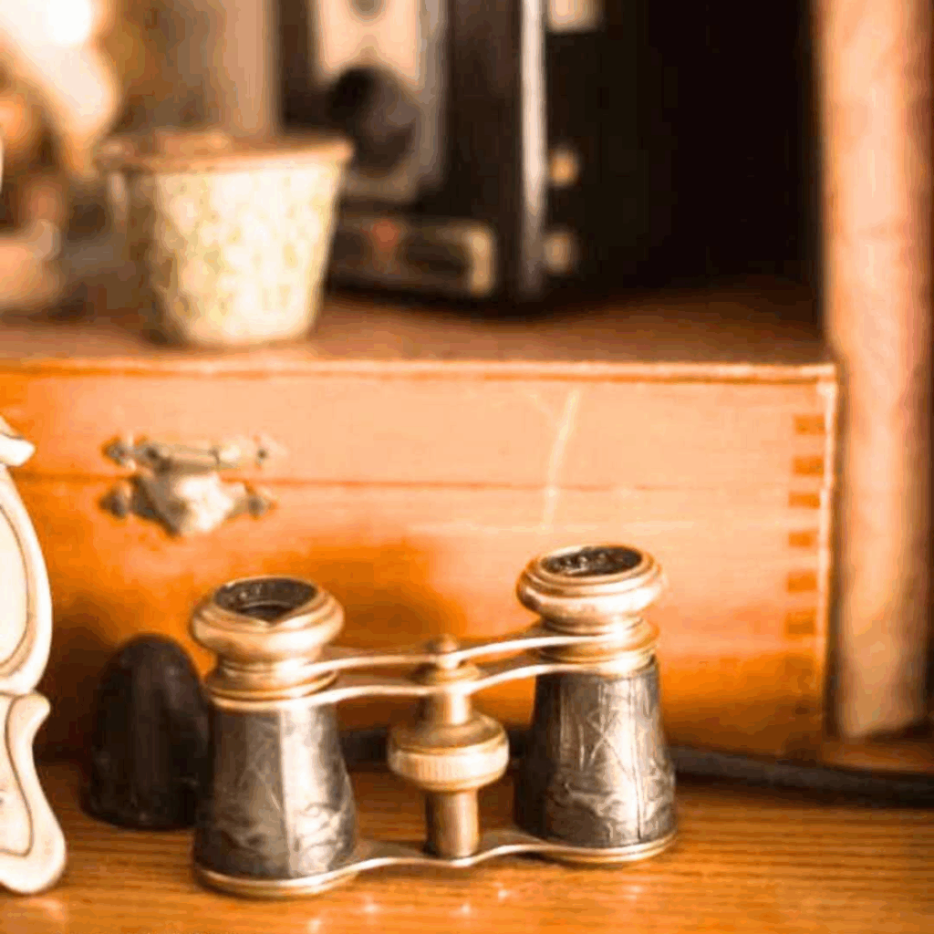 Vintage binoculars glasses with ornate detailing rest on a wooden surface, surrounded by a small ceramic container and a wooden box, evoking nostalgia.