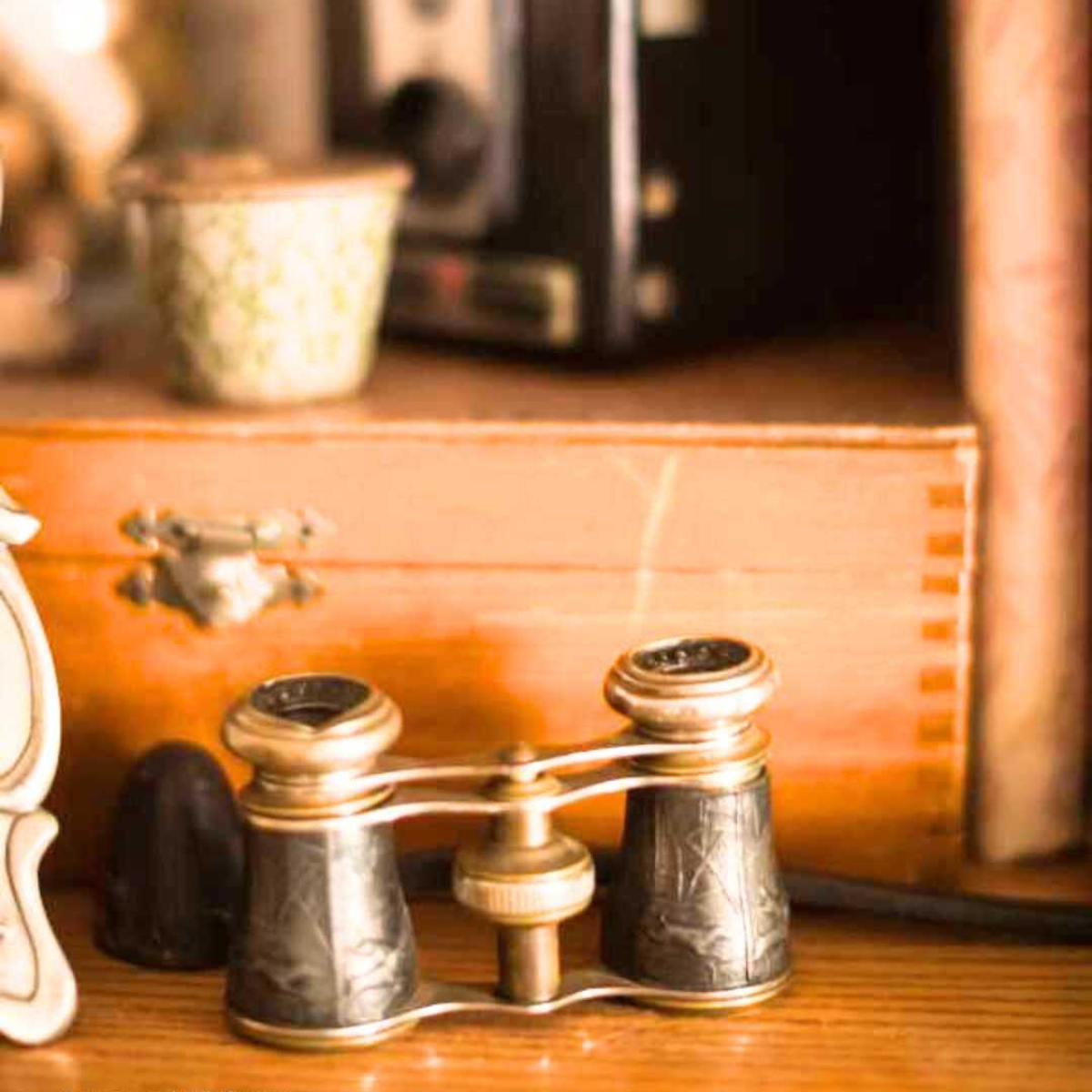 Vintage binoculars glasses with ornate detailing rest on a wooden surface, surrounded by a small ceramic container and a wooden box, evoking nostalgia.