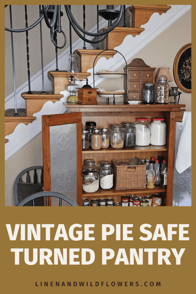 Vintage pie safe repurposed as a pantry under a staircase. Filled with jars and baskets, creating a rustic and cozy atmosphere. Decorative metal doors ajar.