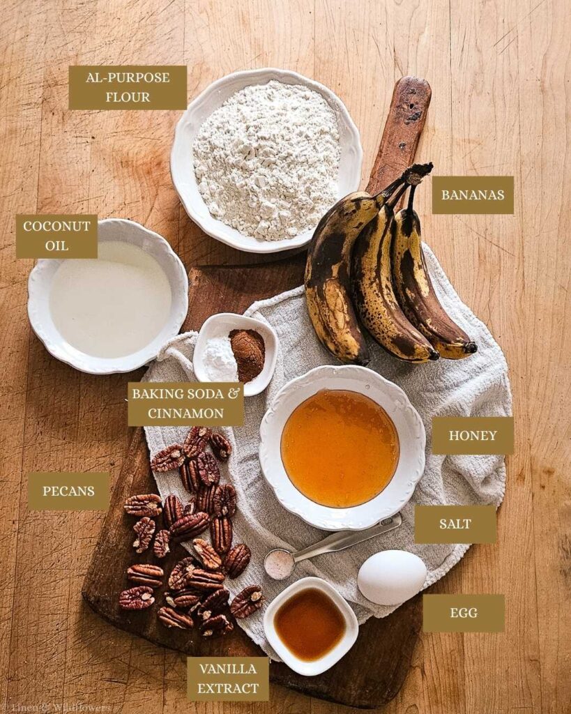 Ingredients on a wooden board for banana bread: ripe bananas, flour, honey, coconut oil, pecans, egg, vanilla, baking soda, cinnamon, salt. Rustic and inviting.