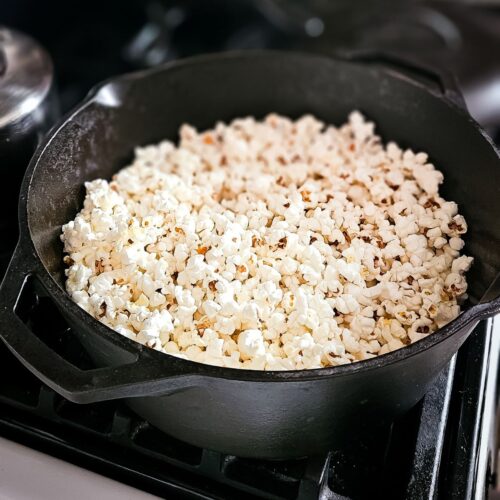 A black cast iron pot filled with freshly popped popcorn sits on a stovetop. The fluffy white popcorn contrasts with the dark pot, creating a cozy, inviting atmosphere.