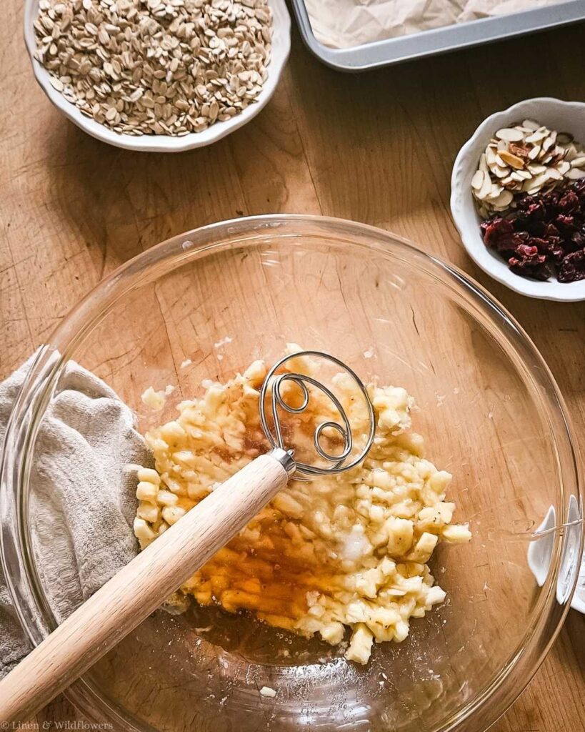A mixing bowl with mashed bananas and vanilla extract on a wooden table. Nearby are bowls of oats, sliced almonds, and dried cranberries, suggesting baking prep.