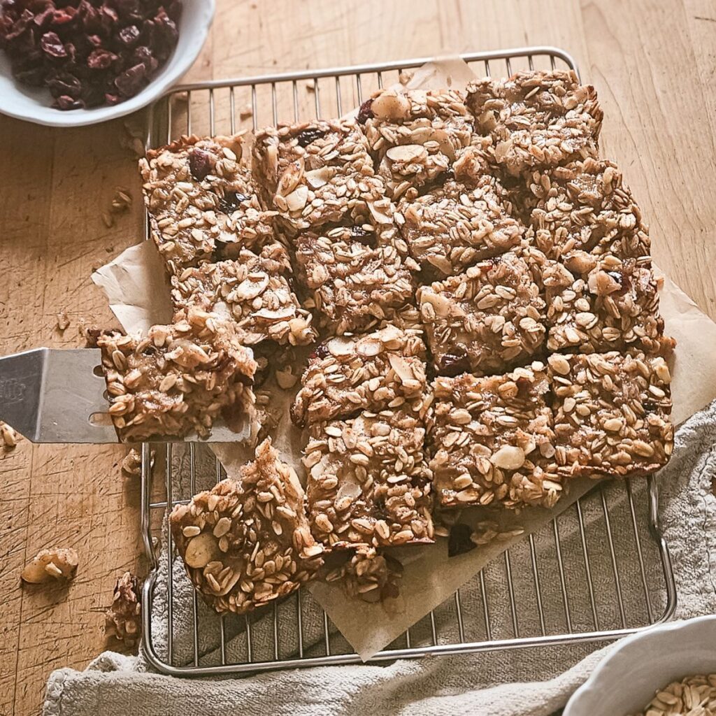 Granola bars with oats and nuts are cut into squares on a cooling rack over parchment paper. A spatula holds one bar, and a bowl of dried cranberries sits nearby.
