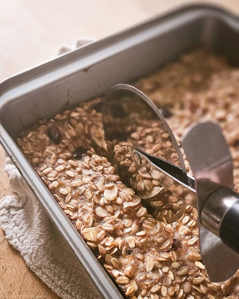 Baked oatmeal in a metal pan is being cut with a pizza cutter. The oats are golden brown, sprinkled with raisins. The setting is warm and inviting.