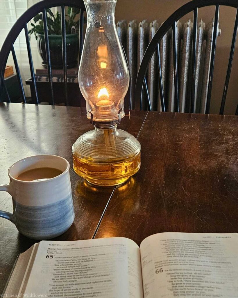 A lit oil lamp casts a warm glow on a wooden table with an open book and a mug of coffee. A cozy, reflective atmosphere prevails.