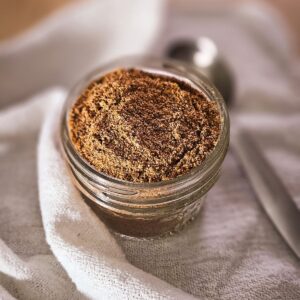 A small glass jar filled with ground spices rests on a textured white cloth. A metal spoon lies beside it, creating a warm, inviting kitchen scene.