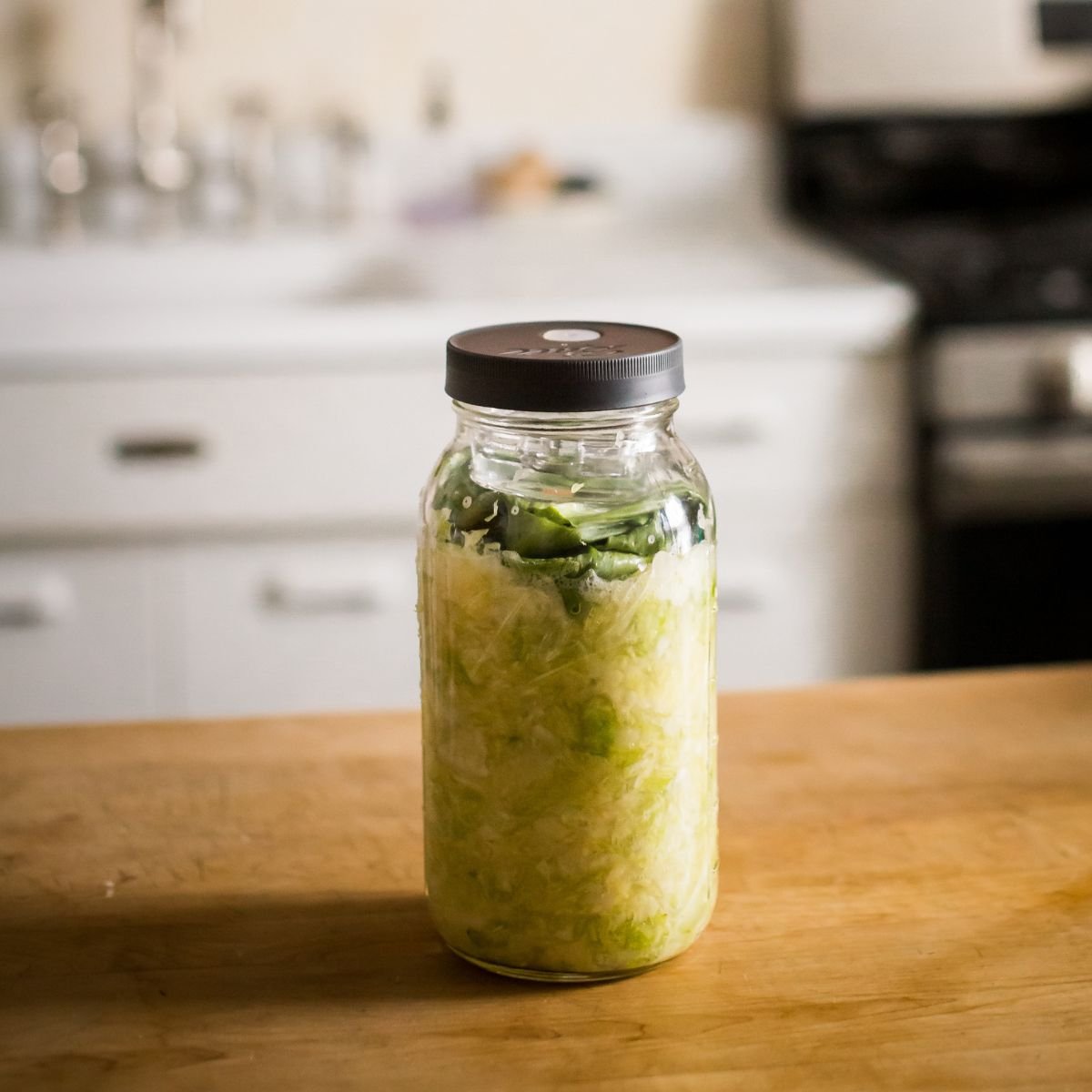 A glass jar on a wooden table contains layered shredded cabbage with leaves on top. Soft kitchen lighting and a blurred stove in the background create a homey feel.