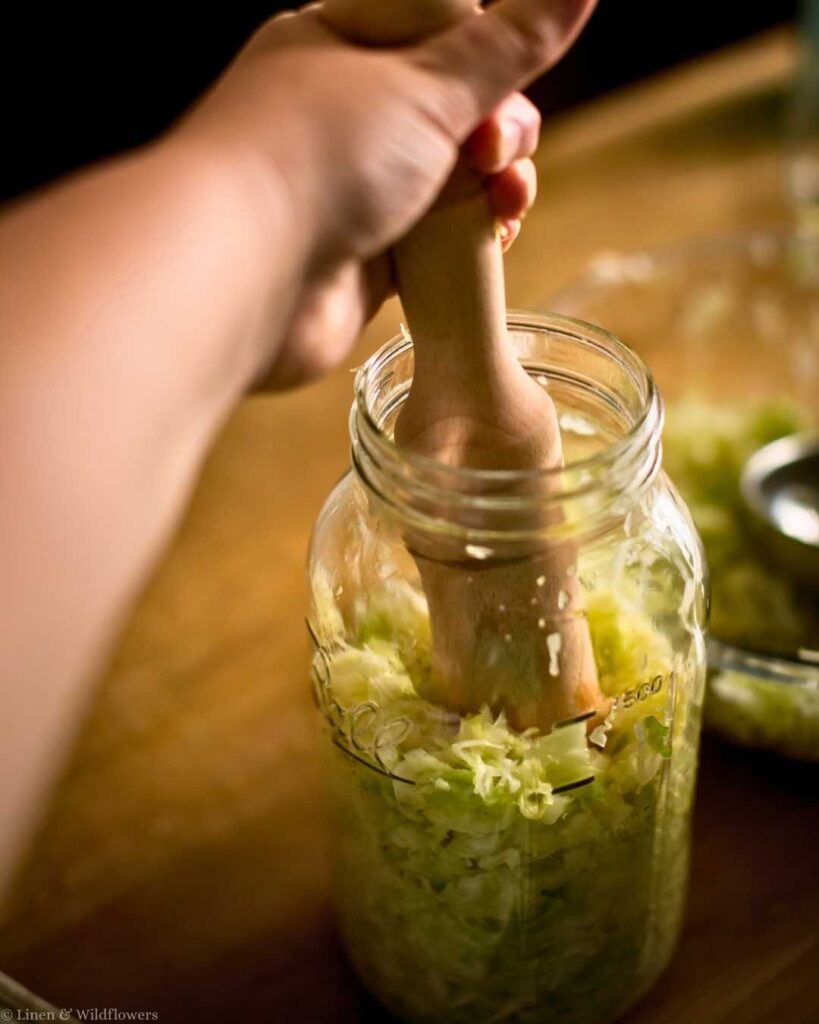 A hand uses a wooden tamping tool to press shredded cabbage into a glass jar for fermentation, set on a wooden surface. The scene suggests homemade sauerkraut preparation.