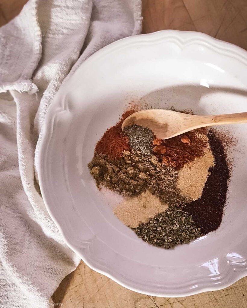 A white plate with assorted spices, including paprika, cumin, and oregano, arranged in sections. A wooden spoon rests on the mix. Soft, earthy tone.