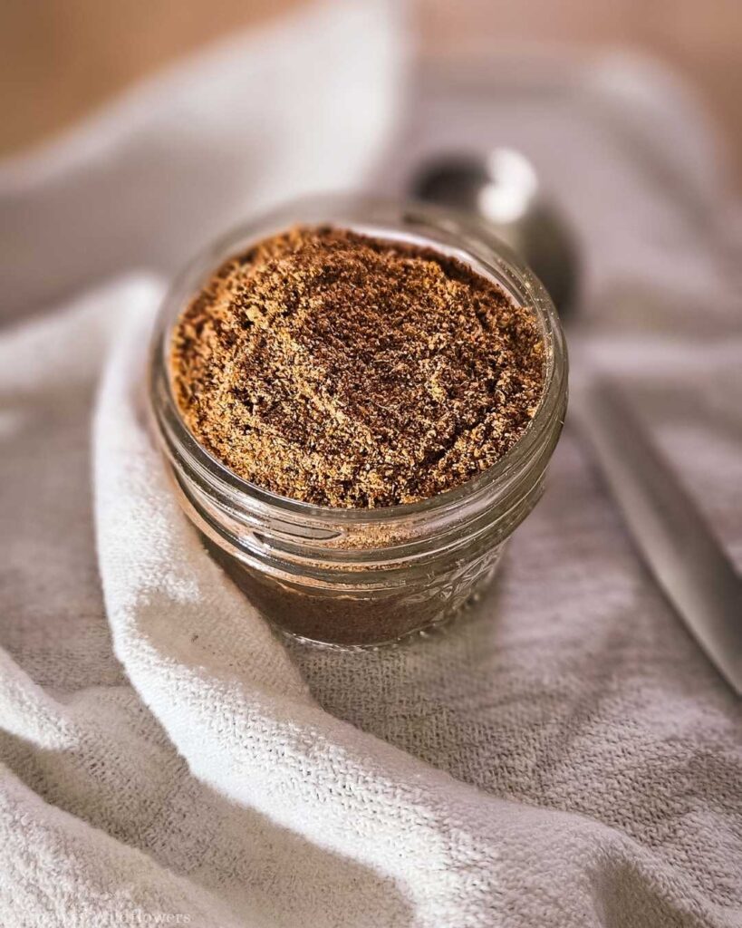 A small glass jar filled with brown sugar sits on a textured white cloth. The setting is softly lit, conveying a warm and cozy atmosphere. A metal spoon is nearby.