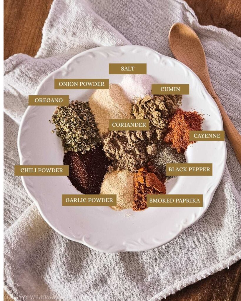 A white plate with assorted spices, including oregano, chili powder, onion powder, salt, coriander, garlic powder, smoked paprika, cumin, black pepper, and cayenne, labeled on a cloth. Wooden spoon beside it. Cozy, warm tones.