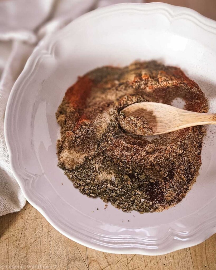 A white bowl holds a blend of herbs and spices, including oregano and paprika, on a wooden surface. A wooden spoon rests in the mix, suggesting preparation.