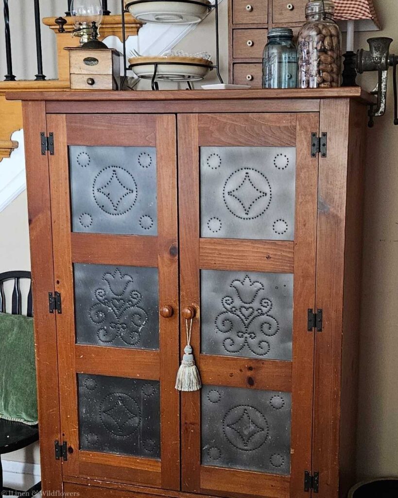 Vintage wooden cabinet with metal paneled doors featuring ornate punched designs. Items, including dishes and jars, are displayed on top, evoking a rustic charm.