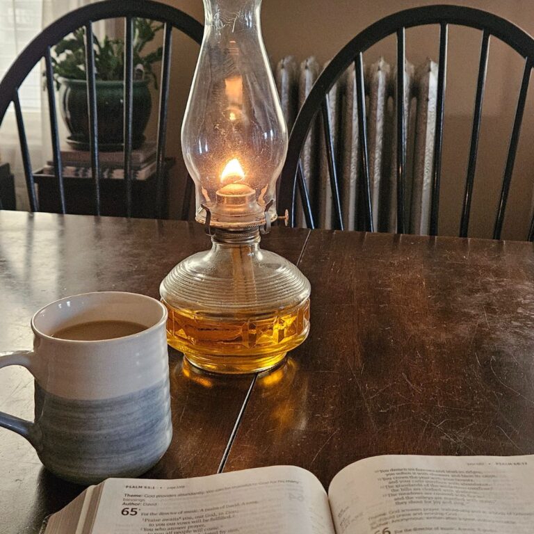 A cozy scene with an open book, a mug of coffee, and a lit oil lamp on a wooden table. The soft lighting creates a warm, peaceful atmosphere.
