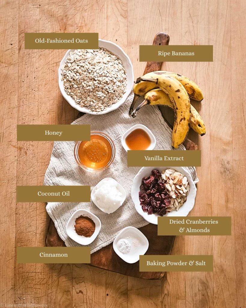 Overhead view of labeled baking ingredients on a wooden board: oats, bananas, honey, vanilla, coconut oil, cranberries, almonds, cinnamon, baking powder, and salt.