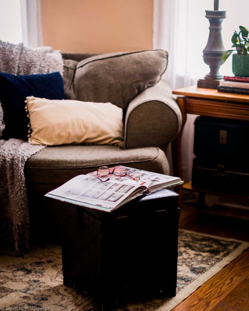 Cozy living room with a gray armchair, soft pillows, and a draped throw. A magazine and glasses rest on a small table, exuding warmth and relaxation.