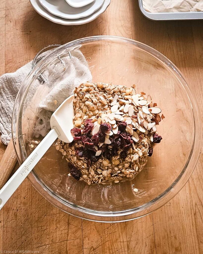 A clear glass bowl on a wooden table contains oats, sliced almonds, and dried cranberries with a white spatula beside it, ready for baking.