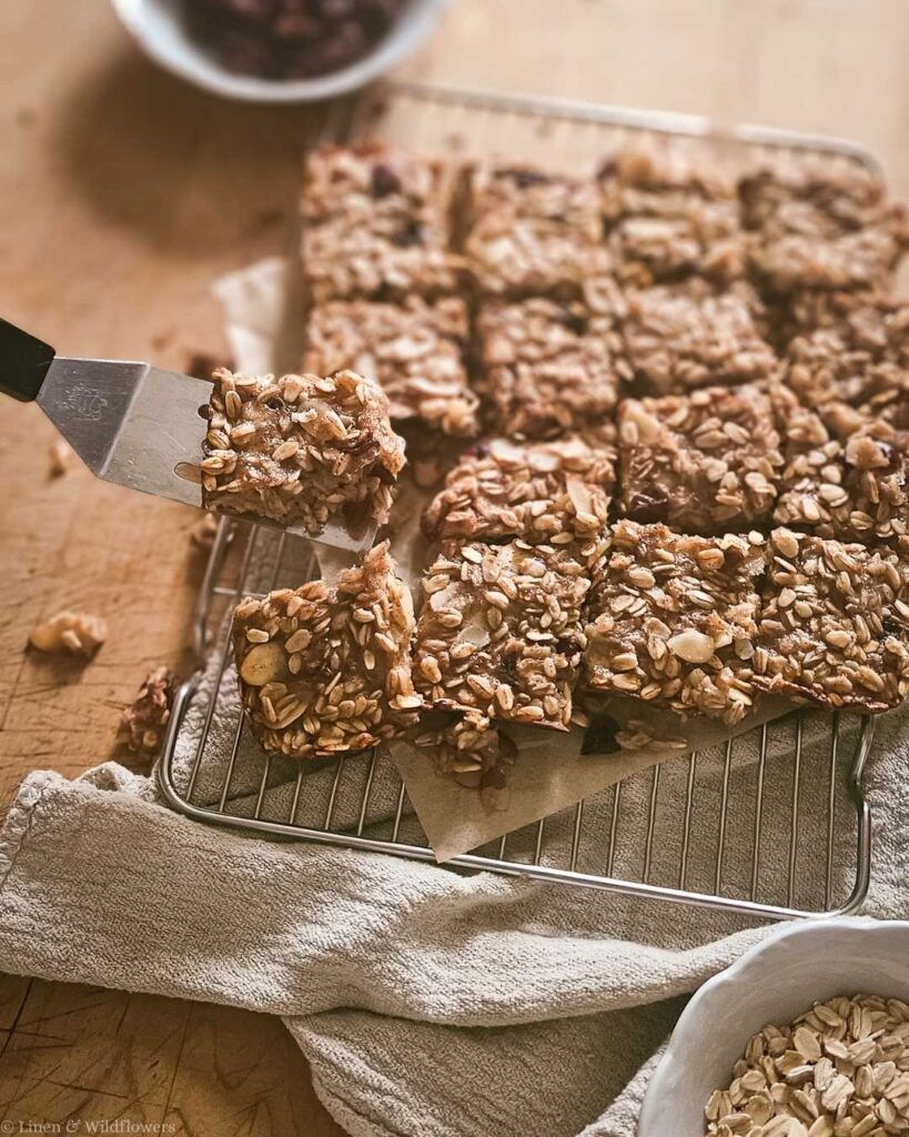 A spatula holds a square oat and nut bar above a grid of similar bars on a cooling rack. A textured cloth and a bowl of oats are nearby. Warm, rustic feel.