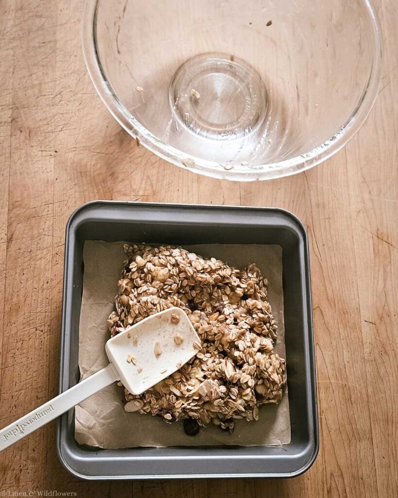 Granola mixture with oats and nuts in a square baking pan with a white spatula on parchment paper. An empty glass bowl is above on a wooden table.