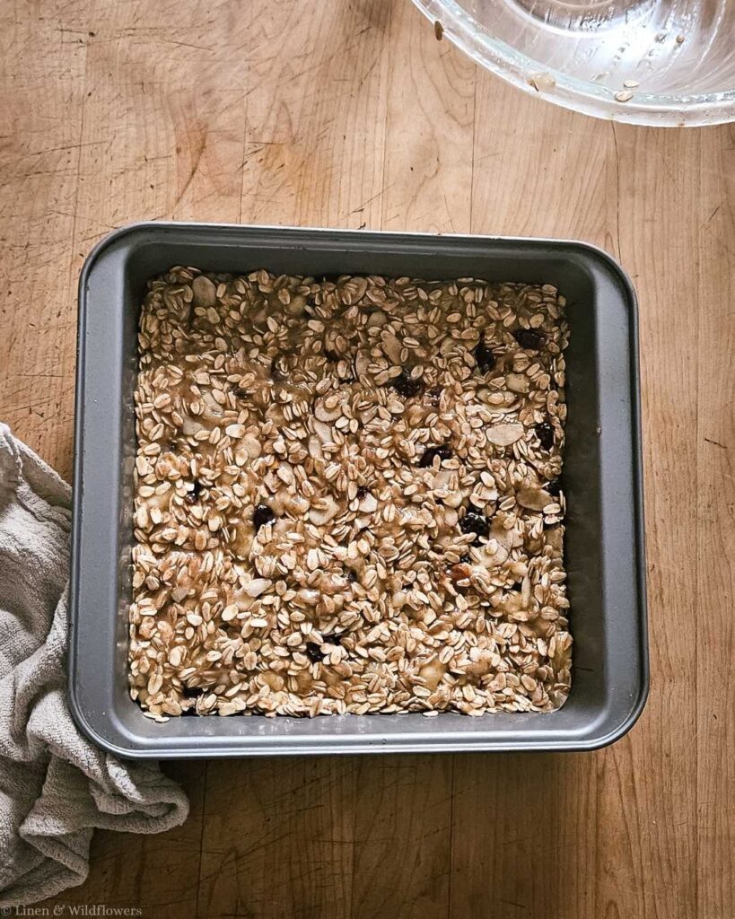 A square baking pan filled with unbaked oatmeal bars, featuring oats and raisins, rests on a wooden surface. A cloth napkin lies nearby, creating a rustic feel.