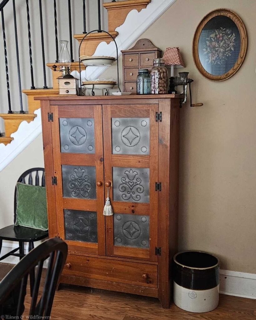 A rustic wooden cabinet with decorative tin panels stands by a staircase. It holds vintage items and jars, with a framed floral picture above, creating a cozy, nostalgic ambiance.