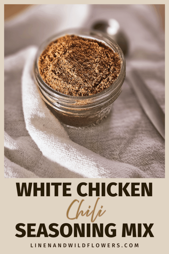 A small glass jar filled with a brown spice blend labeled "White Chicken Chili Seasoning Mix" rests on a white cloth beside a metal spoon.