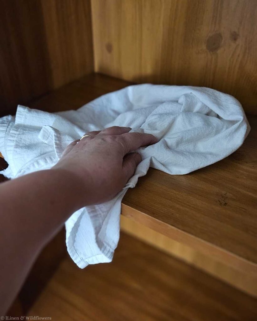 A hand cleaning a wooden shelf with a white cloth, suggesting routine maintenance and tidiness. The wood has a warm tone, conveying a cozy atmosphere.