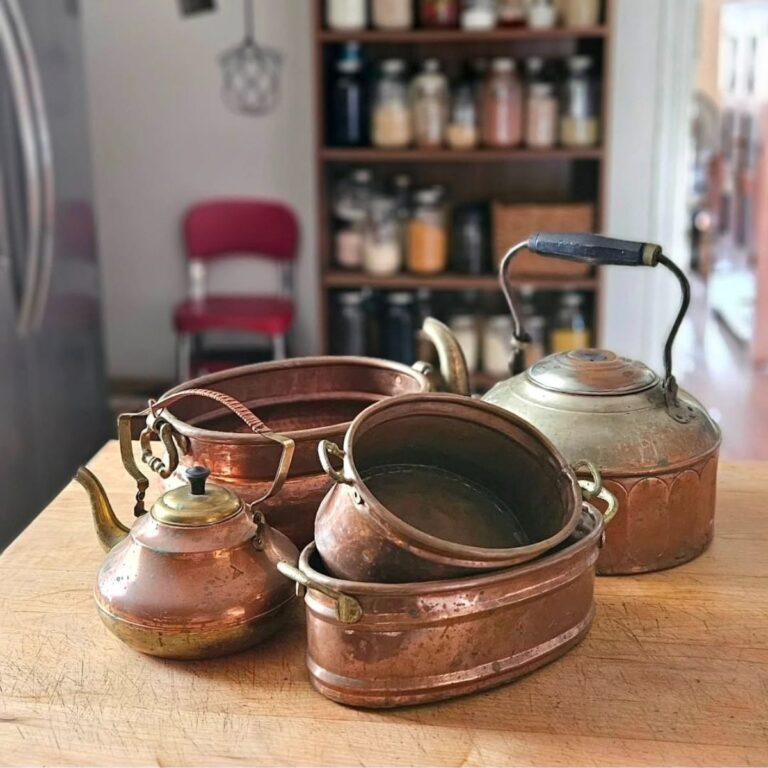 Antique copper kitchenware, including kettles and pots, placed on a wooden countertop. Background features a pink chair and shelves with jars. Cozy ambiance.