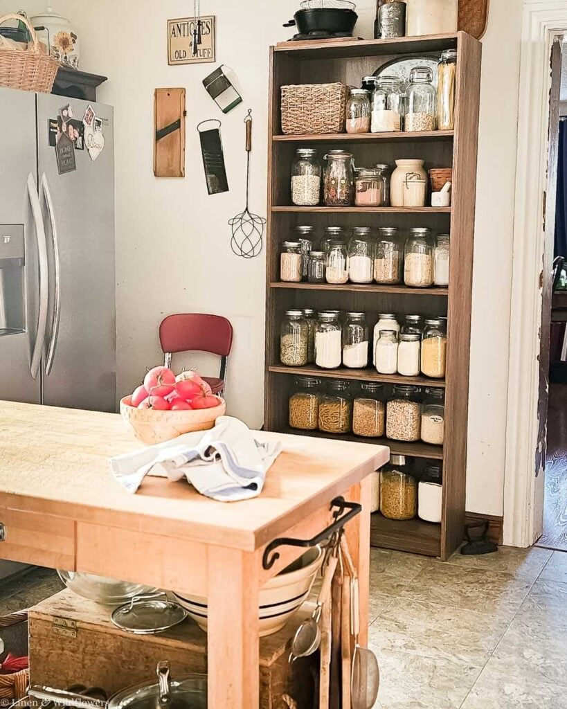 Cozy kitchen with a wooden shelf filled with jars of grains and spices. A wooden island holds a basket of tomatoes and a cloth. Vintage decor evokes warmth.