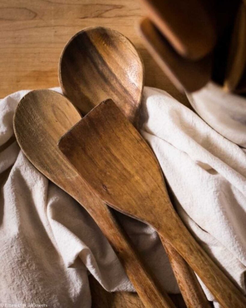 Three rustic wooden cooking utensils rest on a soft, white cloth, set against a warm wooden background. The scene evokes a cozy, homely feel.