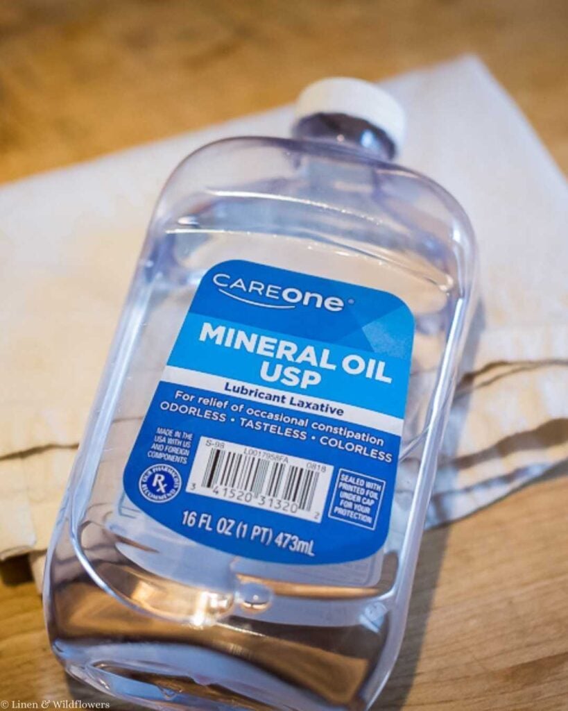A bottle of CareOne Mineral Oil USP is placed on a wooden surface with a cloth in the background. The label notes it as an odorless, tasteless lubricant laxative.