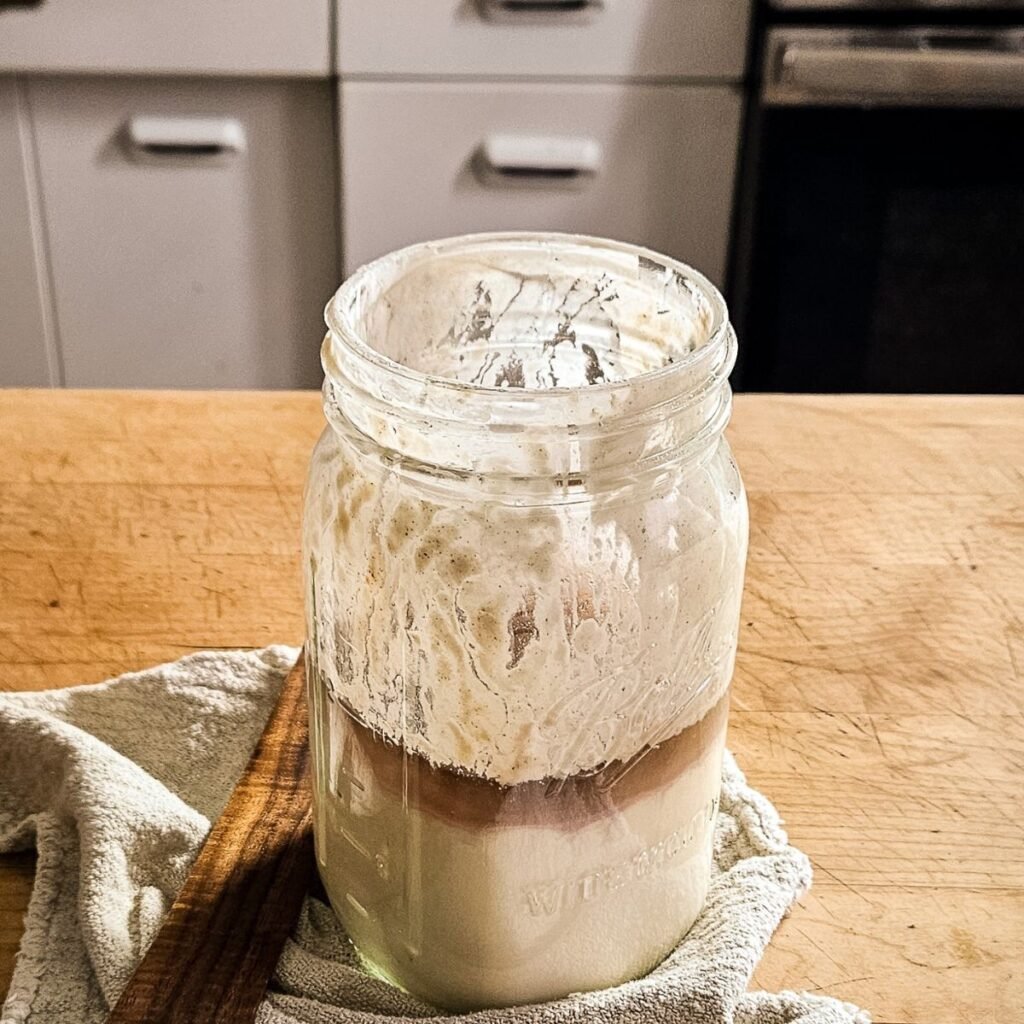 A glass jar containing sourdough starter with visible hooch sits on a wooden countertop, partially wrapped in a cloth, against a kitchen background.