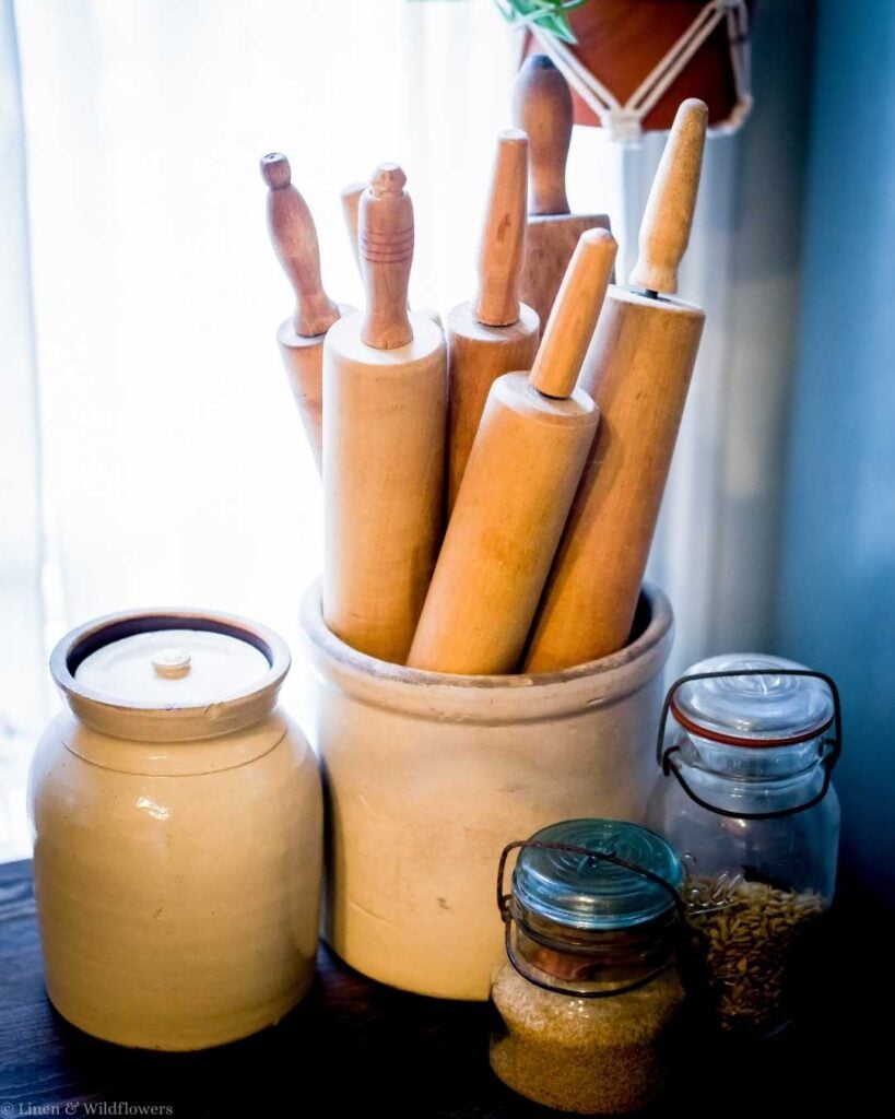A pottery crock holds seven wooden rolling pins on a kitchen counter, flanked by sealed jars and a ceramic container. The scene feels rustic and homely.