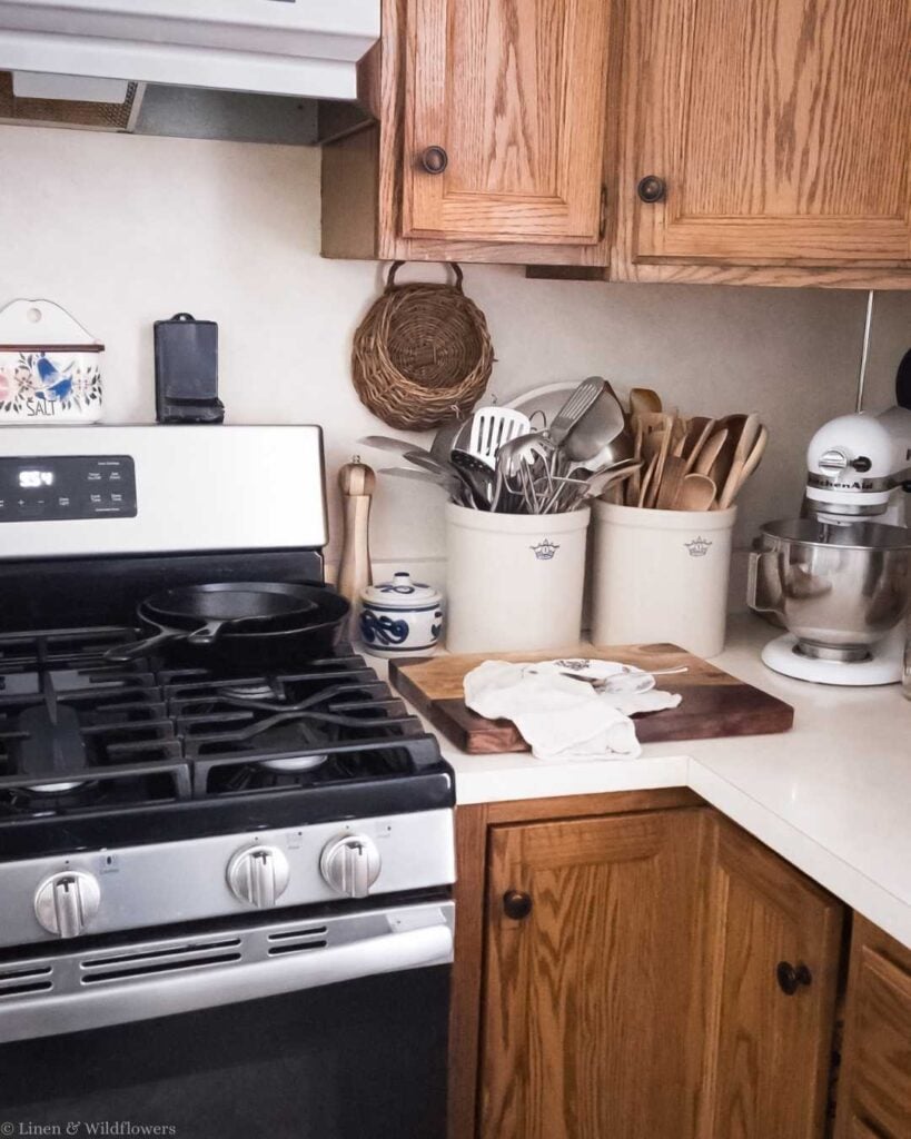 Cozy kitchen corner with wooden cabinets, a stove with a black skillet, and ceramic containers holding utensils; vintage and inviting atmosphere.