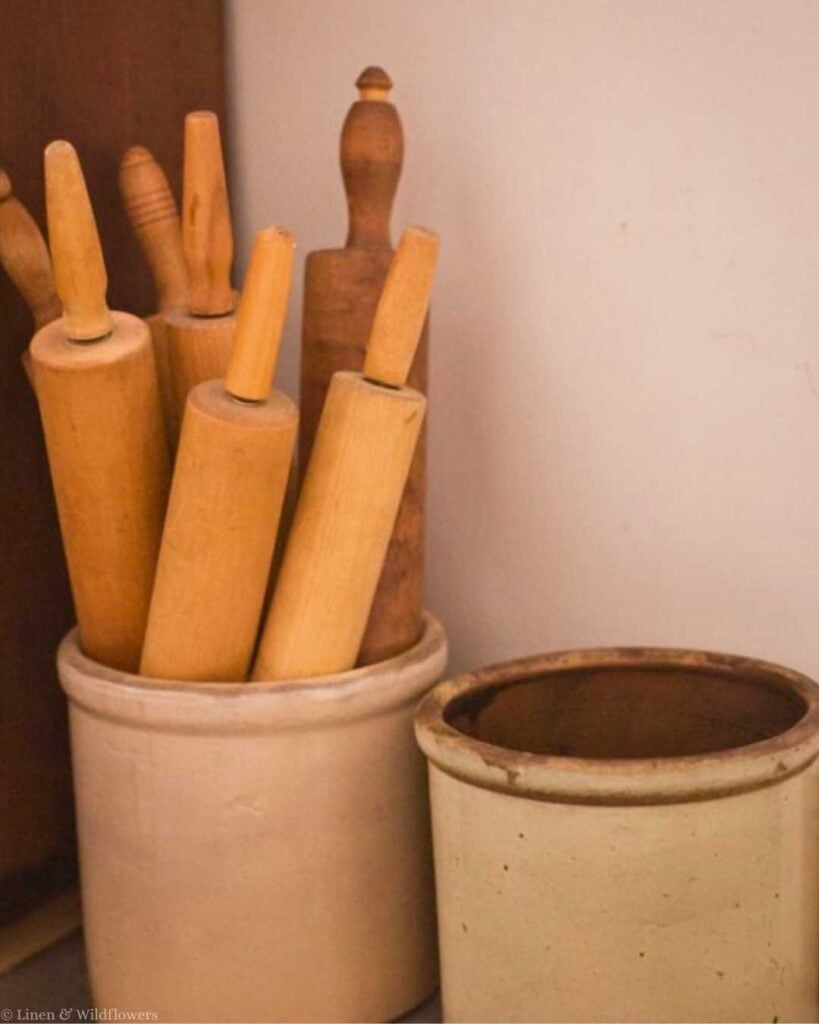 A collection of wooden rolling pins in a cream ceramic jar, next to an empty similar jar, against a soft brown background, creating a rustic, cozy feel.