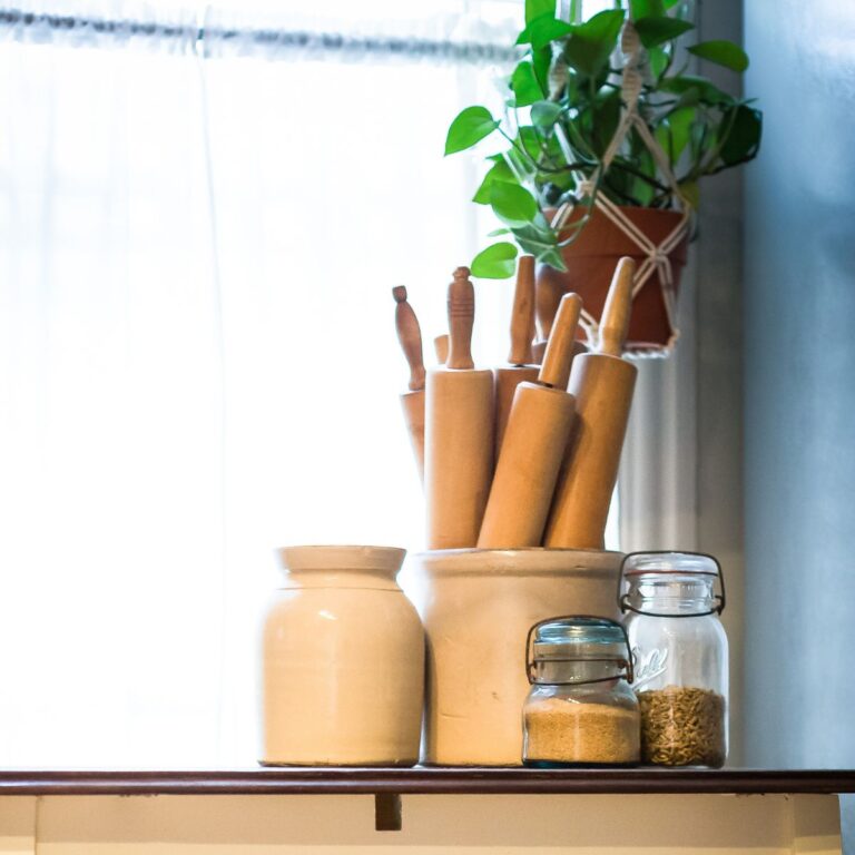 A cozy kitchen windowsill with a rustic feel features a white jar filled with wooden rolling pins, flanked by two glass jars of grains and a hanging plant nearby.