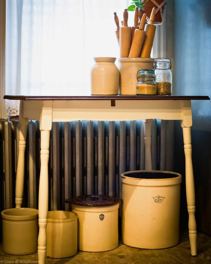 A cozy kitchen scene featuring a wooden table with rolling pins in a crock, a plant, jars, and clay pots. Warm, rustic ambiance.