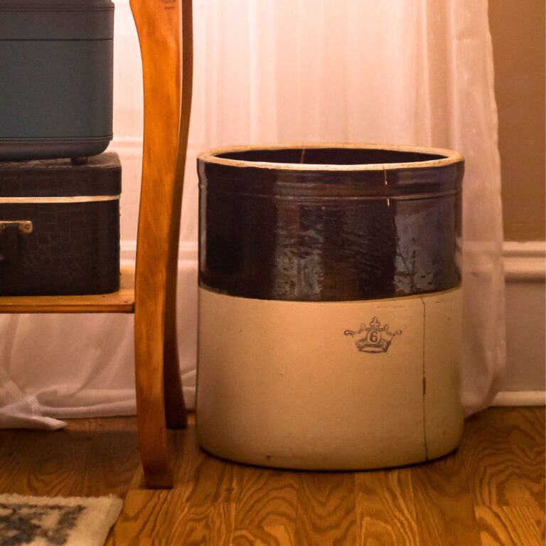 A vintage crock with a dark brown top and cream bottom sits on a wooden floor beside a table with stacked black suitcases, against a sheer curtain backdrop.