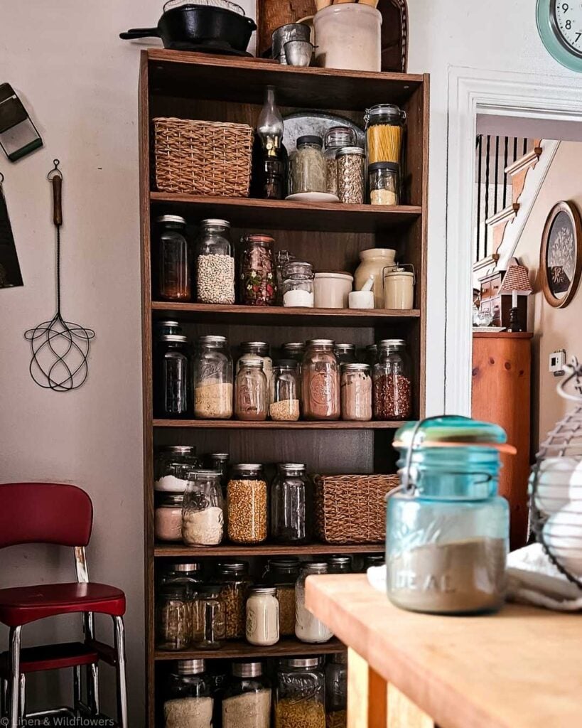A cozy kitchen scene with a wooden shelving unit filled with various jars of grains, spices, and pasta. A red chair is to the left, creating a vintage feel.