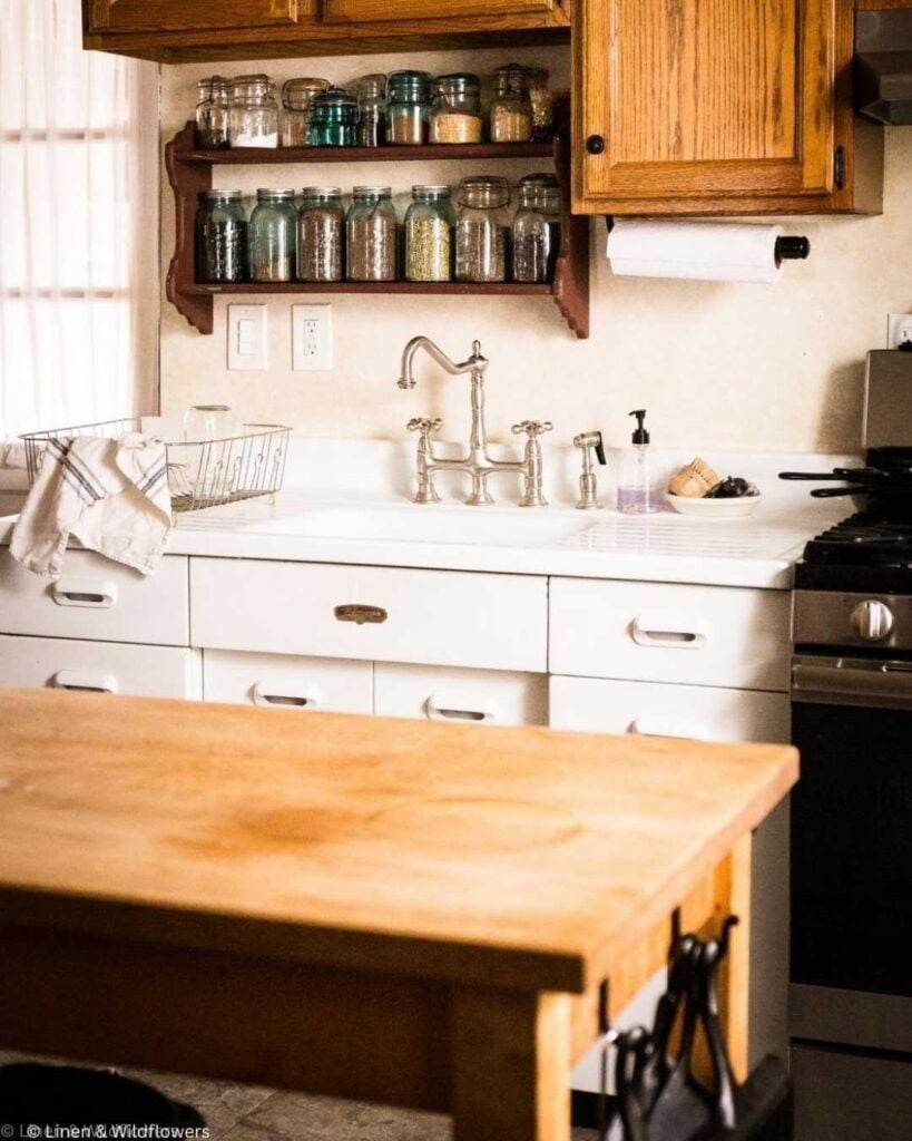 A cozy kitchen with white cabinets and a farmhouse sink. Wooden shelves hold glass jars. A wooden table and a dish rack with a towel complete the scene.