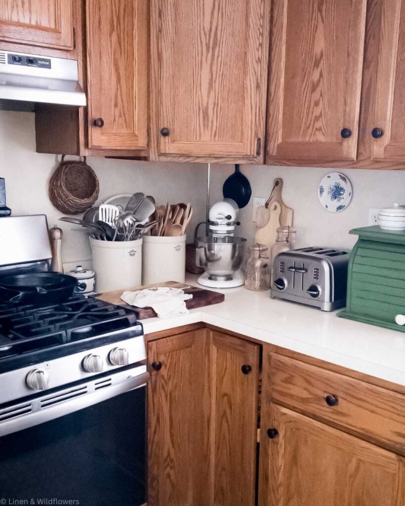 A cozy kitchen corner with wooden cabinets, a stainless steel stove, utensils in ceramic holders, a stand mixer, toaster, and green bread box on white counters.