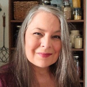 A woman with long gray hair smiles gently in a cozy kitchen. Behind her, shelves display jars and baskets, creating a warm and inviting atmosphere.