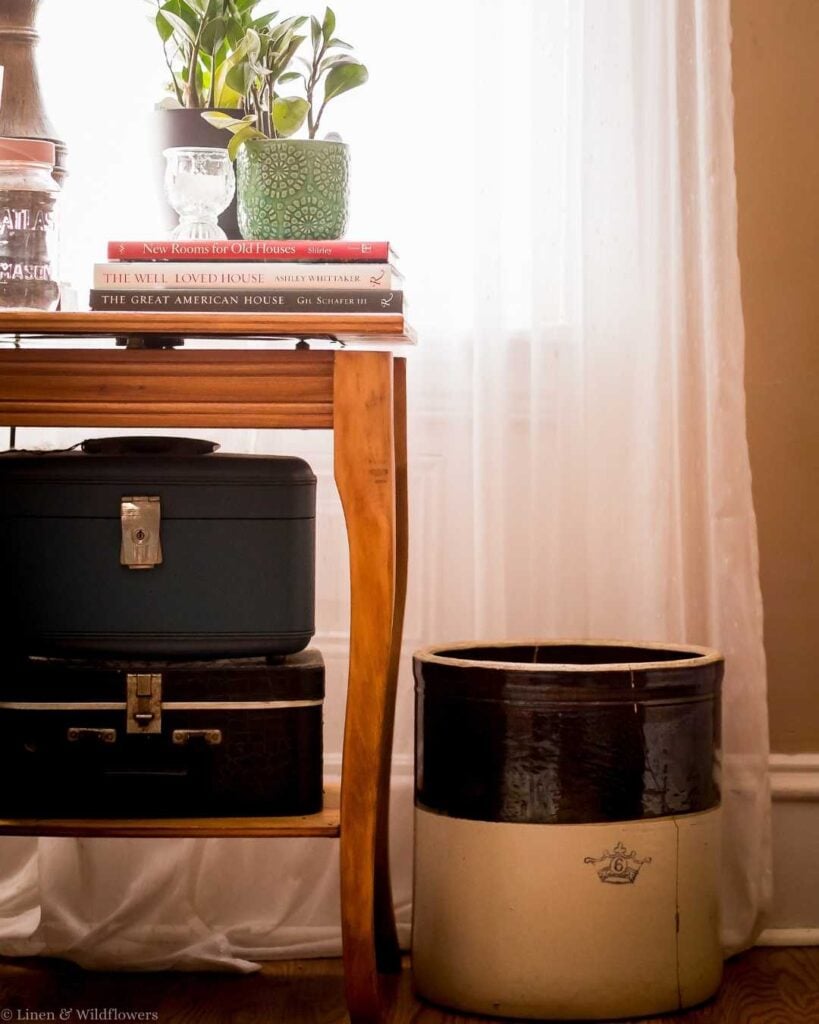 A cozy room corner with a wooden table holding potted plants and stacked books titled "New Rooms for Old Houses." A vintage suitcase and crock sit underneath.