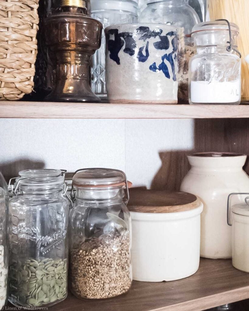 Rustic pantry shelves with glass jars filled with seeds, grains, and a jar of white powder. Ceramic containers and wicker basket add vintage charm.