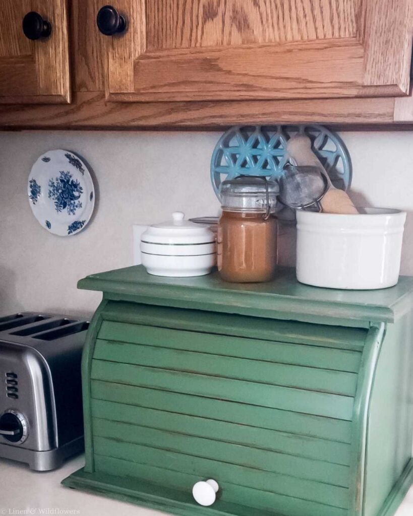 A cozy kitchen nook featuring a vintage green bread box with jars and a tin on top. Wooden cabinets above and a blue floral plate adorn the wall.