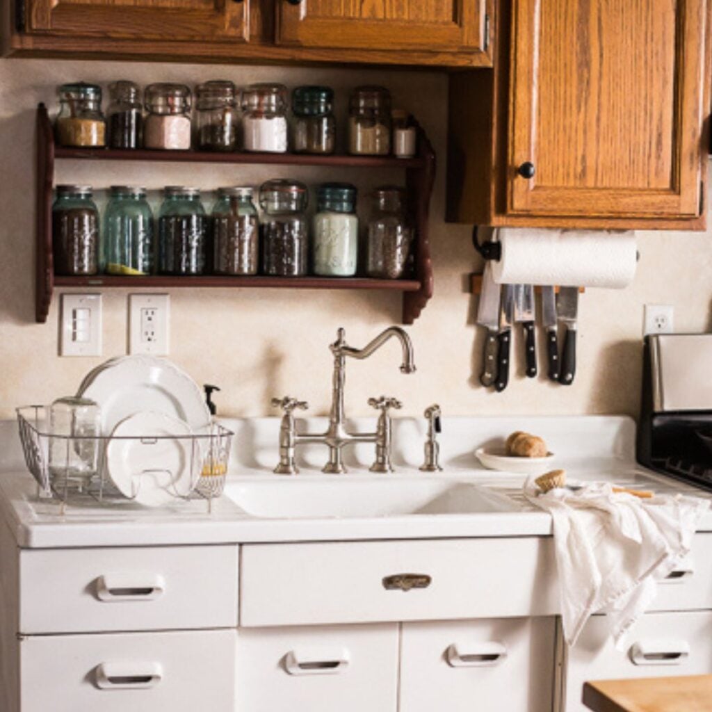 A cozy kitchen with a white vintage-style sink, wooden cabinets, and a shelf of glass jars. Knives hang beside the paper towel holder. Warm and inviting.