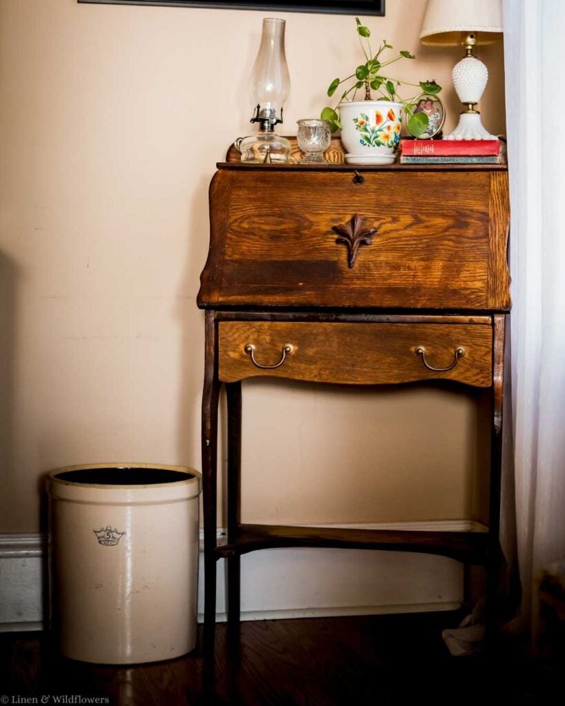 Vintage wooden side table with ornate drawer sits under a cream wall. Oil lamp, potted plant, and books on top. A blush lamp adds warmth.