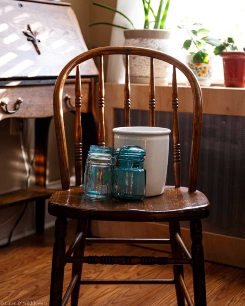 A wooden chair hosts a white crock and two blue and one glass jars, accentuated by warm sunlight. The background features potted plants on a shelf, creating a cozy, rustic ambiance.