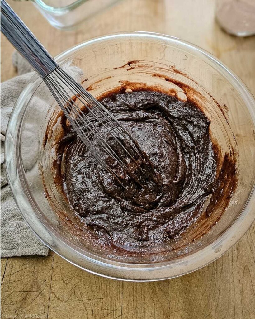 Glass bowl filled with rich chocolate brownie batter being mixed with a whisk. The bowl sits on a wooden surface, evoking a cozy baking atmosphere.