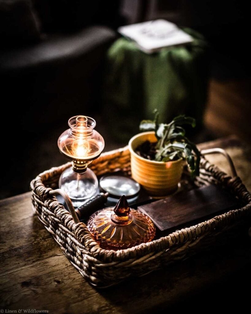 A cozy basket on a wooden table holds a lit oil lamp, a small potted plant, and decorative items. The warm light creates an inviting, rustic ambiance.